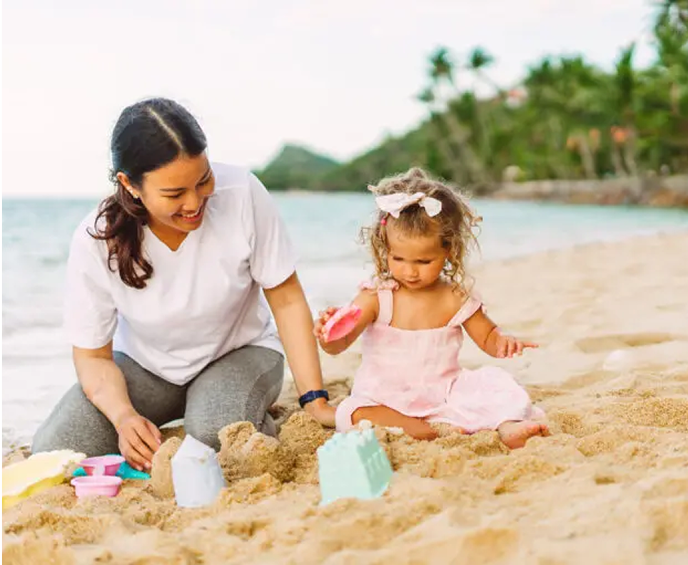Picture → Nannimoon nanny helping a toddler build sandcastles on a sunny beach in Thailand