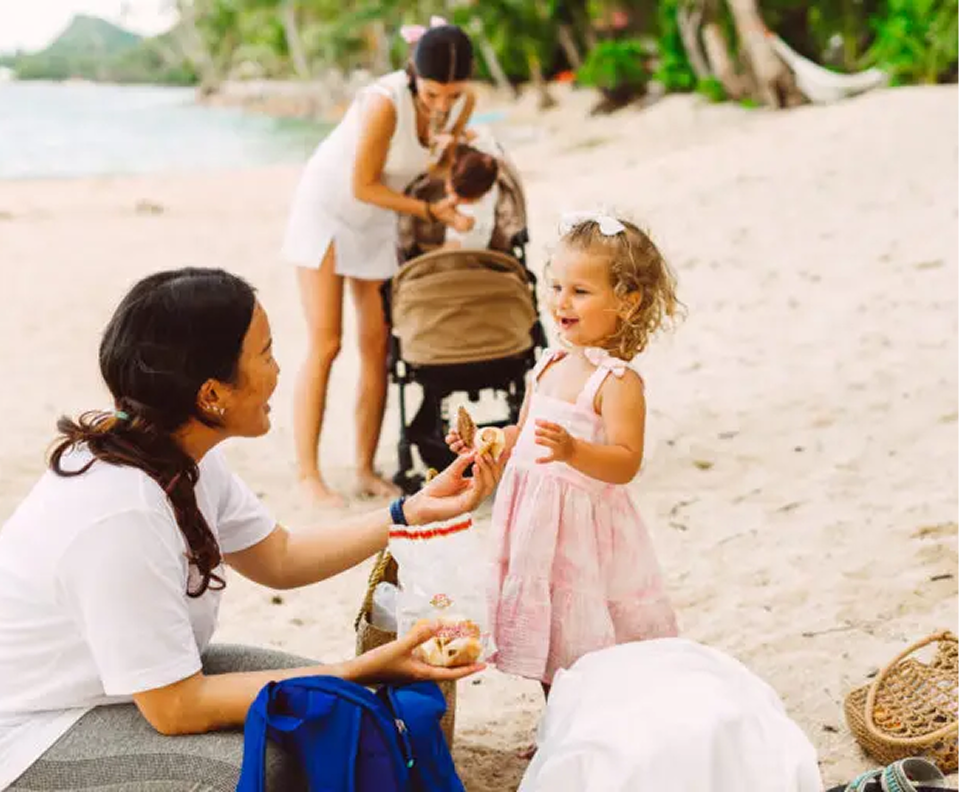 Picture → Nannimoon nanny sharing snacks with a child while a family enjoys the beach in Thailand